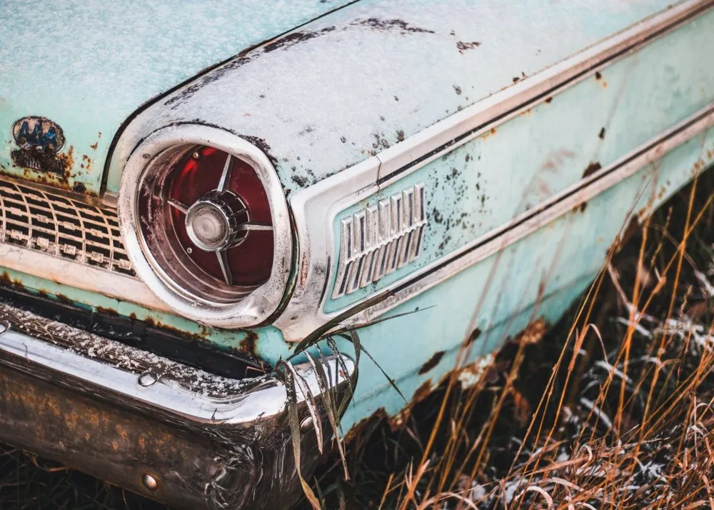 Close-up of a rusted classic car’s headlight and bumper, paint flaking off pale blue metal